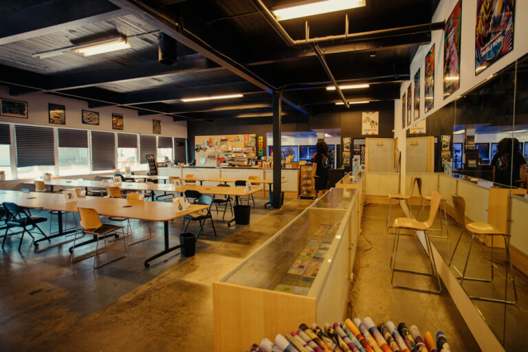 A spacious game store features long tables with chairs, a glass display case, and colorful posters on the walls. Various board games and collectibles are visible. The room has a warm, inviting atmosphere with dim lighting.