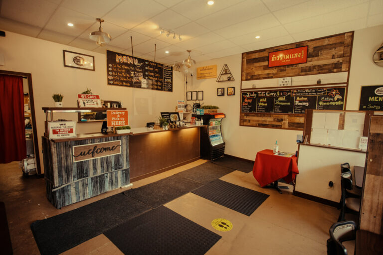 A cozy restaurant interior with a counter displaying menus and signs. A bulletin board shows specials, and small tables are set for dining. Warm lighting and decorations create a welcoming atmosphere.