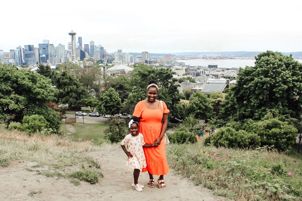 A woman and a child wearing bright dresses stand on a grassy hill with a scenic view of a city skyline in the background, featuring the Space Needle and surrounding buildings. Trees and open spaces are in the foreground.