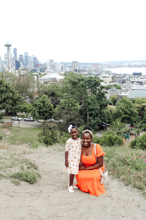 A woman in an orange dress kneels next to a young girl in a floral dress with a white bow, smiling on a grassy hill. The Seattle skyline, including the Space Needle, and trees are visible in the background.