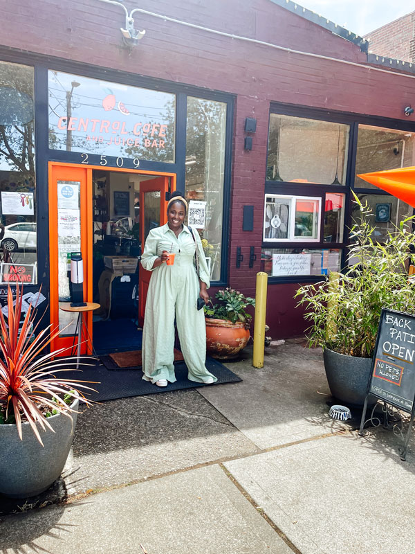 A person in a light green outfit stands outside the entrance of Central Cafe. They're holding a drink and smiling. The cafe has a red brick exterior with potted plants and a sign indicating it's also a juice bar.