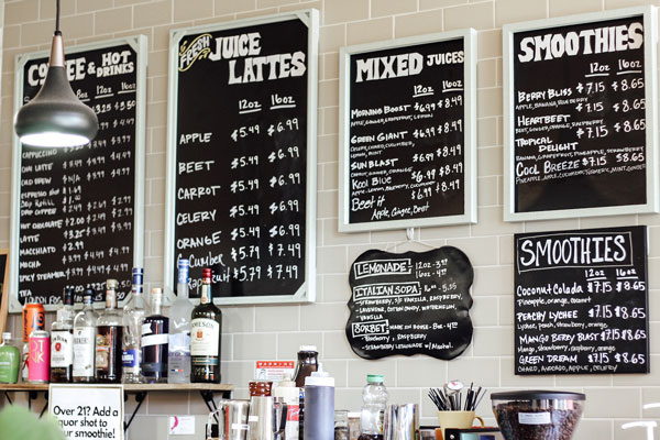 A cafe menu board displays various drinks including coffee, juices, lattes, mixed juices, and smoothies with prices. Bottles are visible on the counter beneath the boards. A pendant light hangs above the area.