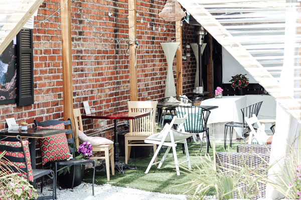 A cozy outdoor patio with a variety of chairs and tables set on artificial turf. The space features brick walls, wooden beams, and several colorful cushions. Potted plants add a touch of greenery, while sunlight filters through the semi-transparent roof.