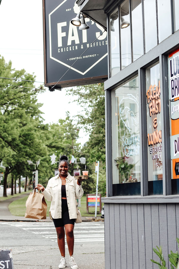 A person in a light jacket and shorts smiles while carrying takeout bags and cups, walking past a restaurant called Fat’s Chicken & Waffles in an urban setting with greenery and street signs.