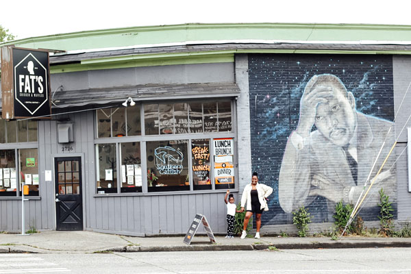 A woman and child stand outside a gray building with a mural of a person holding their head. The storefront has signage for Fat's Chicken and Waffles. The windows display various food and drink-related posters.