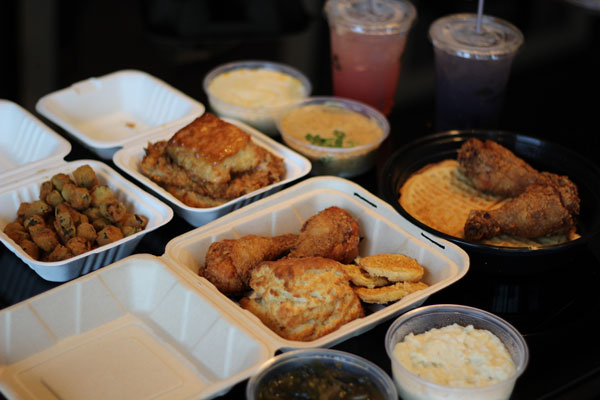 A spread of Southern comfort food including fried chicken, waffles, biscuits, fried okra, mashed potatoes, and greens in takeout containers, accompanied by three drinks in plastic cups.