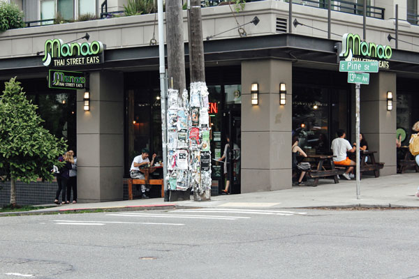 A street corner view of a restaurant named "Manoa Thai Street Eats" with outdoor seating. People are dining outside. There are lamp posts covered with flyers nearby, and street signs for E Pine St and 17th Ave are visible.