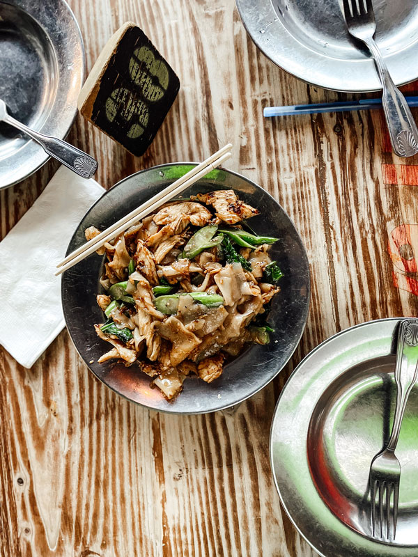 A dish of stir-fried noodles with vegetables is served on a metal plate, accompanied by chopsticks. Additional empty plates and utensils are arranged on a wooden table. A napkin is also visible beside the dish.