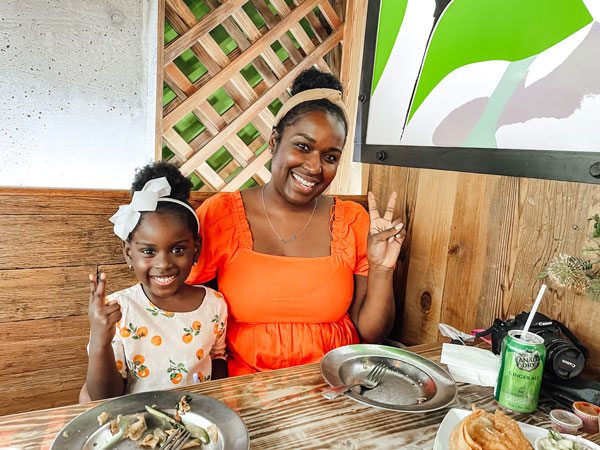 A woman and a young girl sit at a wooden table in a restaurant, both smiling and making peace signs. The woman wears an orange dress, and the girl has a white bow in her hair and a patterned dress. Plates and a green soda can are on the table.