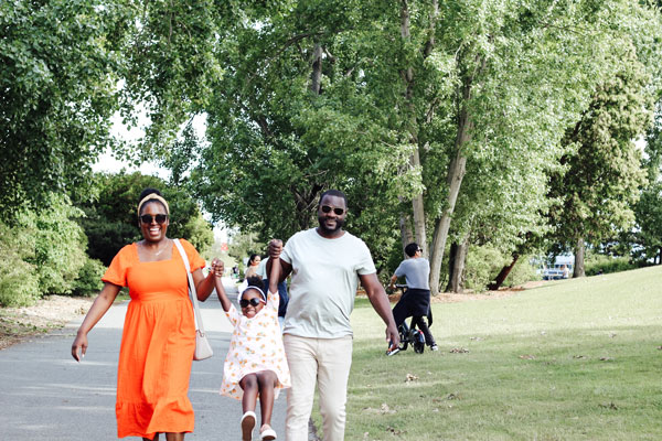 A smiling family of three walks outdoors. The mother in an orange dress and the father in a light shirt hold their daughter’s hands as she swings in a floral dress, wearing sunglasses. They are surrounded by lush green trees and grass.