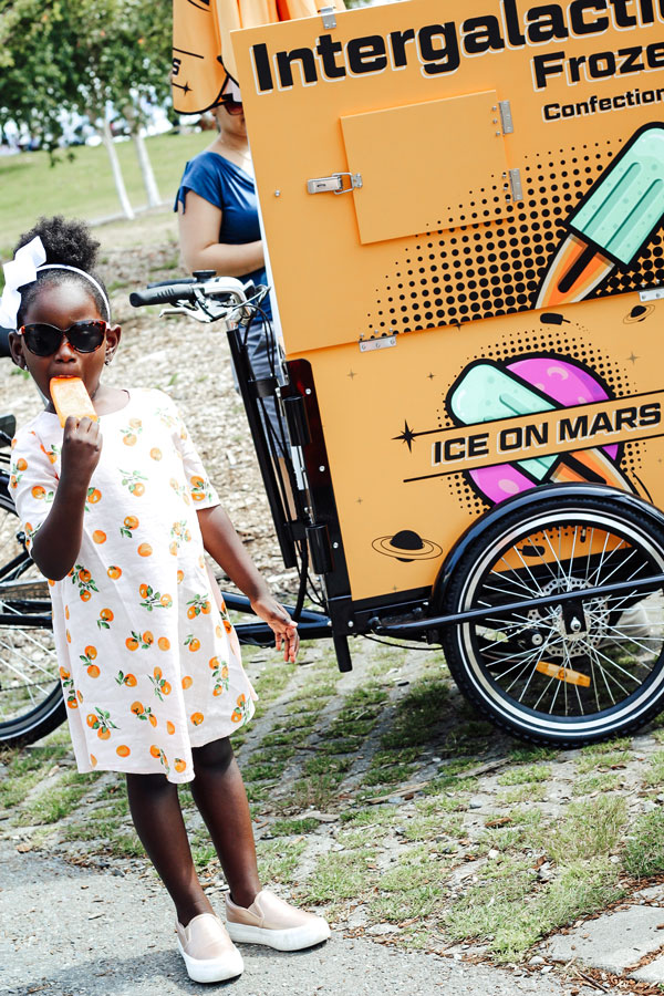 A child wearing a white dress with orange patterns and sunglasses enjoys an orange popsicle. She stands near a bright orange ice cream cart labeled "Intergalactic Frozen Confections" with illustrations of popsicles and "Ice on Mars.