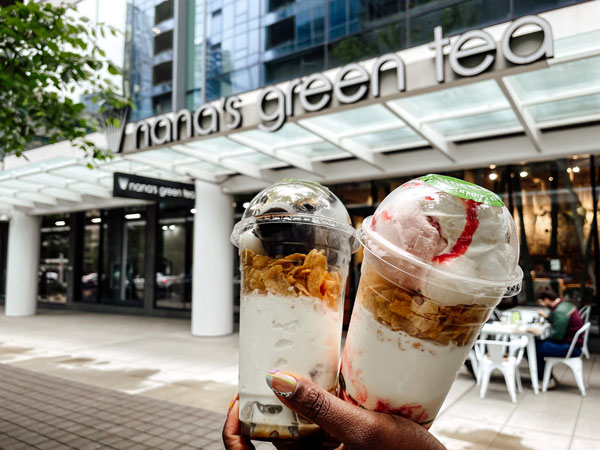 Two hands hold up two parfait cups with layers of yogurt, granola, and fruit toppings in front of a storefront with the sign "nana's green tea." The background shows an outdoor seating area and modern buildings.