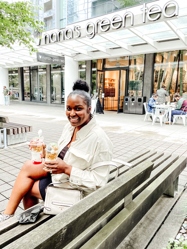 A person sitting on a bench outside nana's green tea cafe, smiling and holding two cups of desserts. The cafe exterior and people seated at tables are visible in the background.