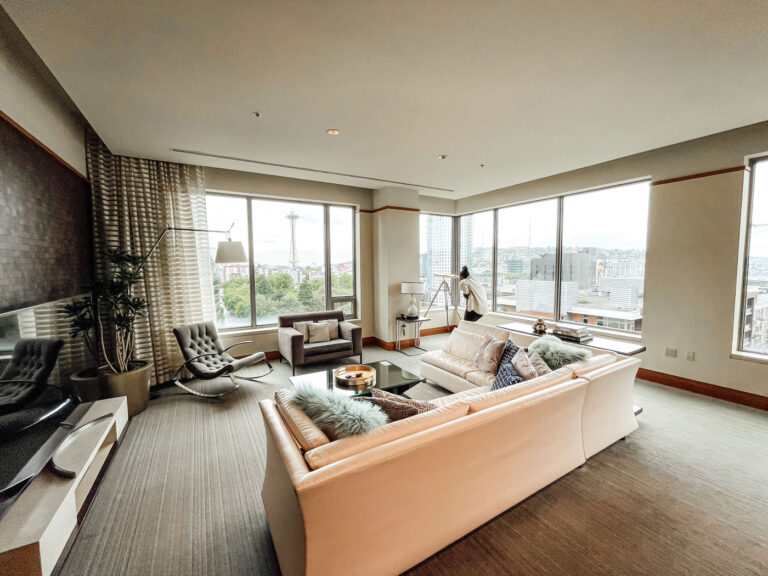 A modern living room with a white sectional sofa facing a flat-screen TV. Large windows offer a view of an urban landscape. The room features a telescope, armchairs, a coffee table, and decorative plants. Neutral tones create a calm atmosphere.