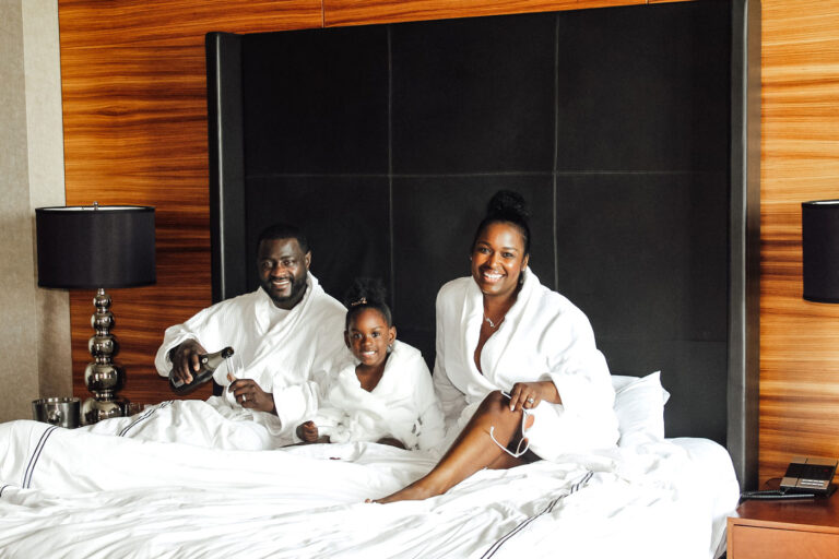 A smiling family of three, wearing white robes, sits on a bed in a stylish room with wooden walls. A man pours a drink, a young girl sits between him and a woman, and two bedside lamps frame the scene.