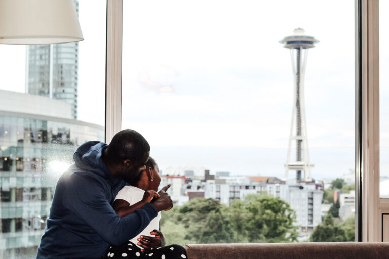 A couple sits by a large window, overlooking the Seattle skyline with the Space Needle prominently in view. They are embracing and looking out at the cityscape.