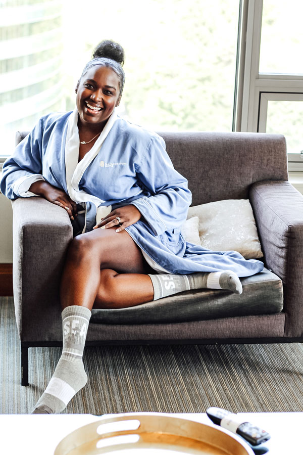 A person wearing a blue bathrobe sits comfortably on a sofa by a window, smiling. They have gray socks with white letters and are holding a mobile phone. A light-colored pillow is beside them on the sofa. A round wooden tray is in the foreground.