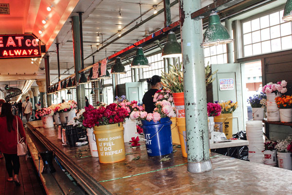A bustling flower market with colorful bouquets in buckets displayed along the counter. Hanging lights illuminate the area, and signs are visible above. A person is arranging flowers amidst the vibrant setting.