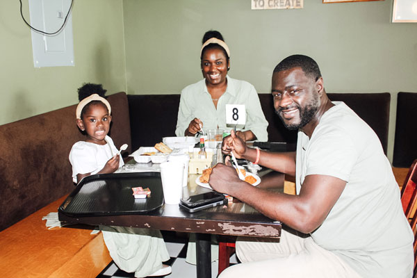 A family of three, two adults and a child, sit at a restaurant table with food. They are smiling and appear to be enjoying their meal. The table has a number 8 sign. The setting is cozy, with a dark brown bench and green walls.