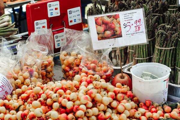 A market stall displaying bags and piles of cherries with a price sign reading "$5.99 per lb." In the background, bundles of asparagus are arranged upright. Payment options are displayed on a red box behind the produce.