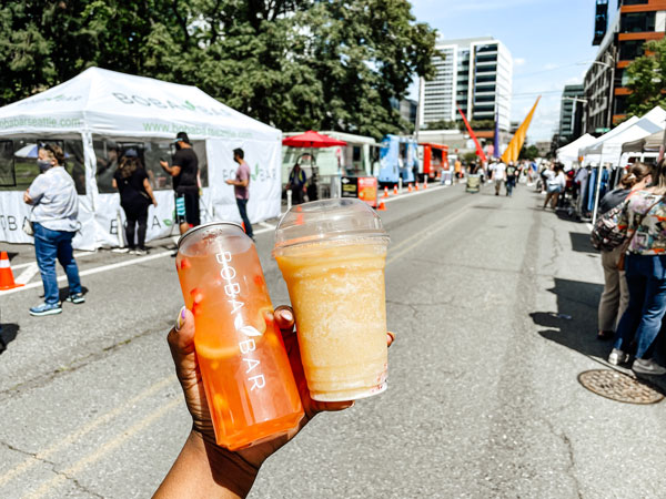 A person holds two drinks on a sunny street lined with festival tents and people. One drink is orange with fruit, and the other is a light yellow smoothie. The scene suggests a lively outdoor market or fair.