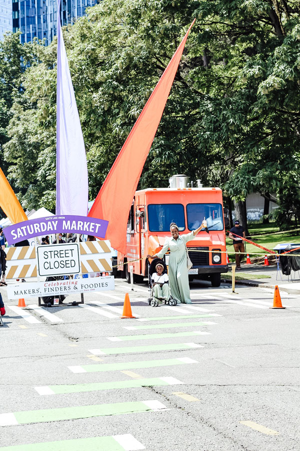 A person in a green dress poses energetically with a child in a stroller at a street closed for a Saturday market. An orange food truck is in the background, surrounded by trees and colorful flags.