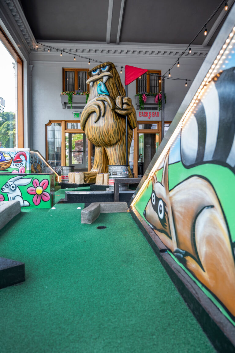 A vibrant indoor mini-golf course with colorful murals featuring cartoon animals. A large statue resembling a fur-covered creature holds a red flag. Strings of lights hang from the ceiling, adding to the playful atmosphere.