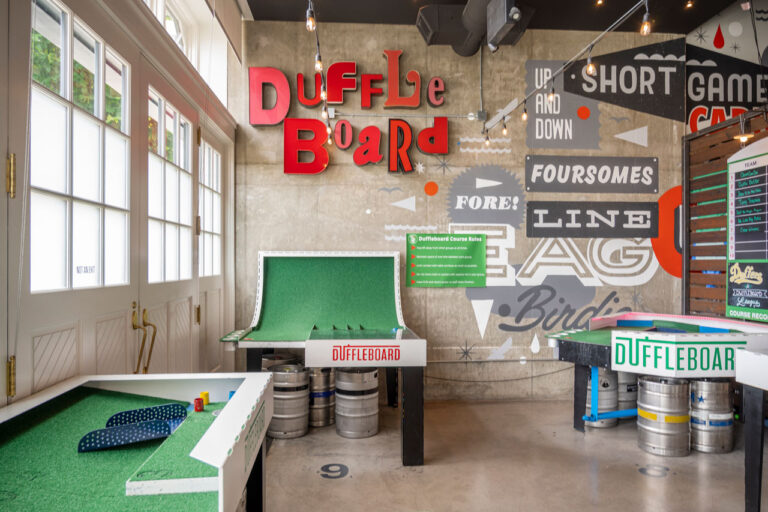 A room featuring duffleboard games with green felt surfaces on top of metal kegs. The walls are adorned with colorful signage and graphics. String lights hang from the ceiling, and large windows allow natural light to illuminate the space.