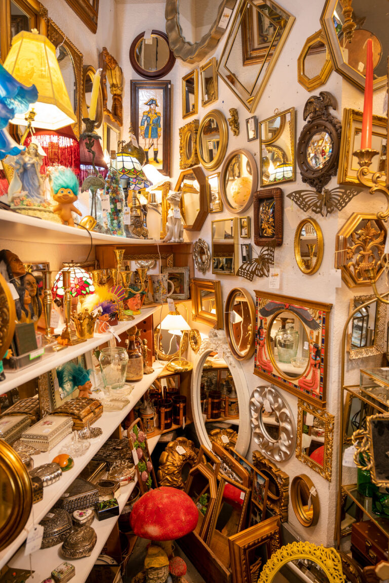 A cluttered, narrow vintage shop filled with various mirrors and picture frames on the walls. Shelves are stacked with eclectic items, including lamps, figurines, and decorative objects. Warm lighting enhances the cozy atmosphere.