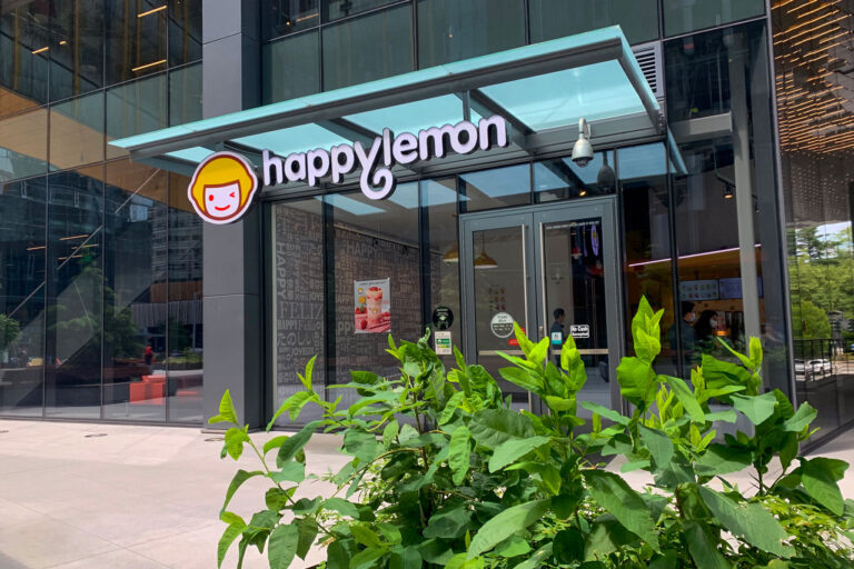 Exterior of a Happy Lemon café with a glass facade. The entrance has a bright sign with a logo of a smiling lemon-headed character. In the foreground, green plants frame the modern, urban setting.