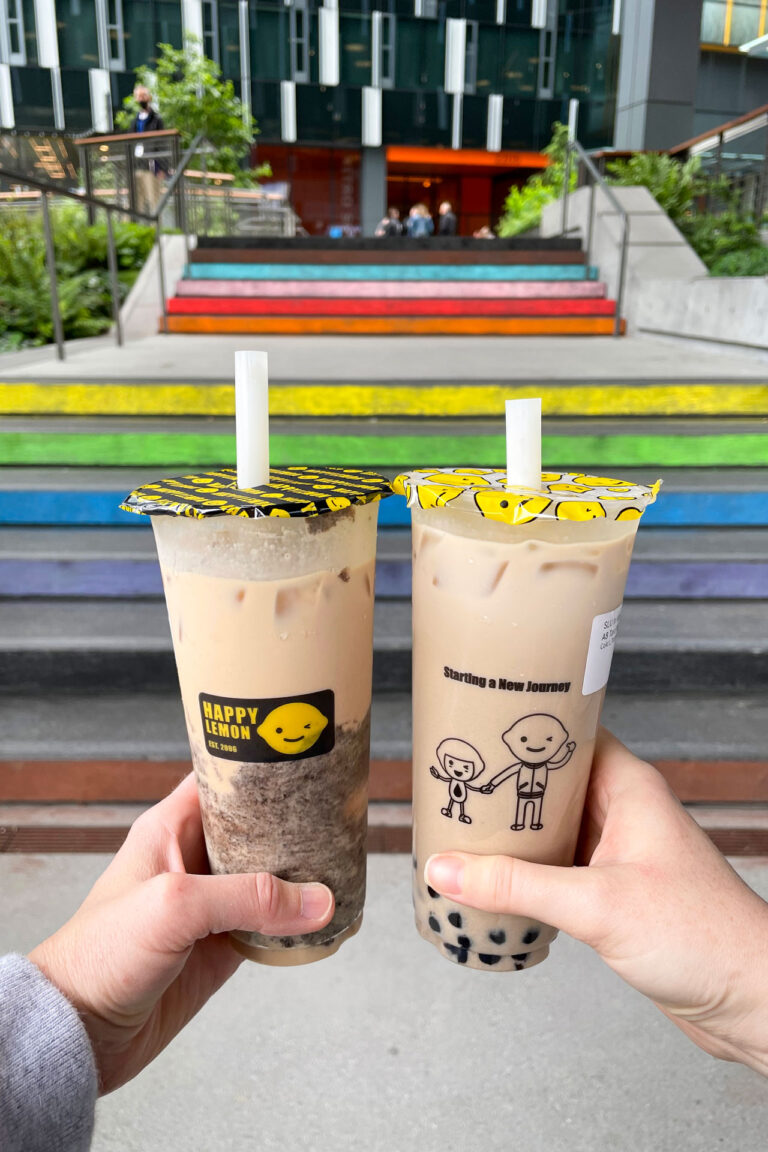 Two hands holding bubble tea cups with colorful lids in front of rainbow-colored steps. The background features a modern building and greenery.