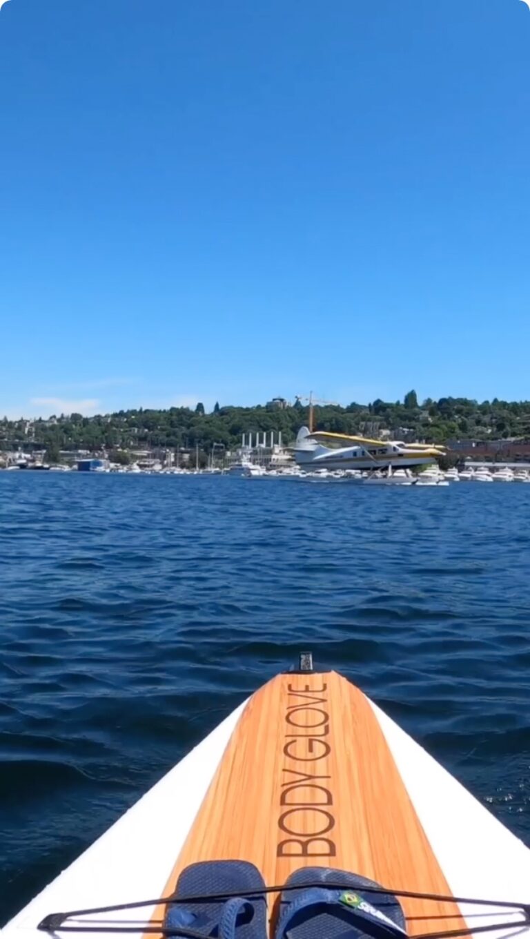 View from a paddleboard on a sunny day showing a blue lake with a seaplane landing in the distance. The paddleboard has "Body Glove" written on it. Forested hills and boats are visible under a clear blue sky.