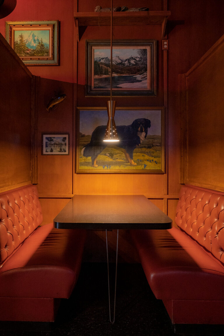 A cozy restaurant booth with red tufted seating and a dark wood table. Warm lighting from a pendant lamp above illuminates the space. The walls are decorated with landscape and animal paintings in wooden frames.