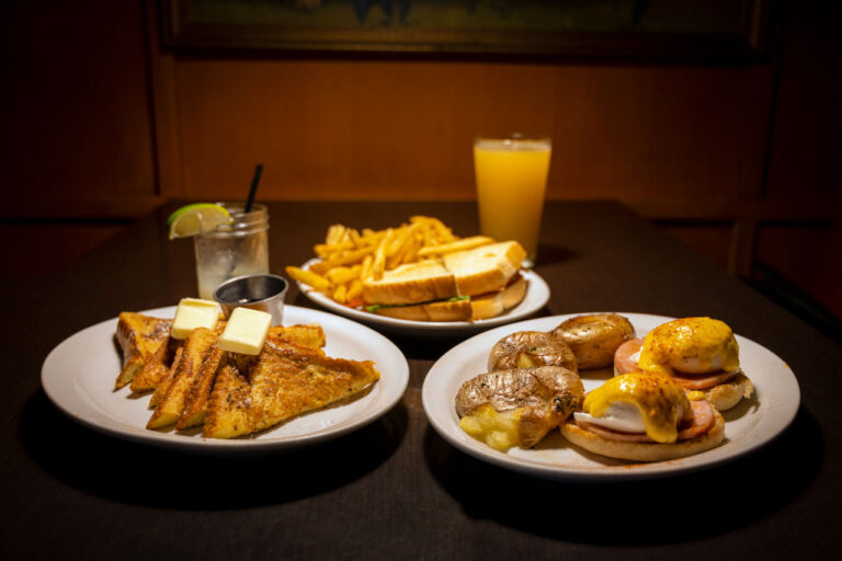 A table with plates of French toast topped with butter, a sandwich with fries, and eggs Benedict with potatoes. Drinks include a glass of orange juice and a cocktail with a lime wedge.