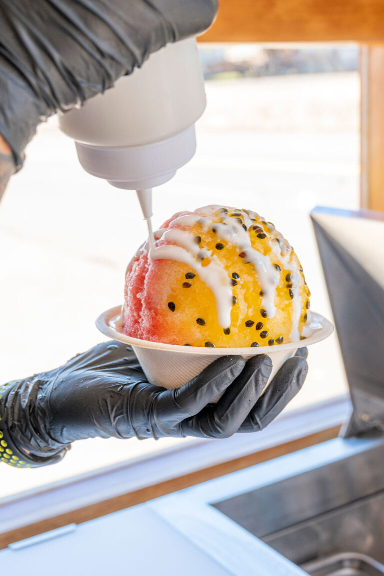 A person wearing black gloves is holding a bowl of shaved ice with red and yellow sections, topped with seeds. White syrup is being drizzled over the ice from a bottle. The scene is set in front of a window with sunlight casting shadows.