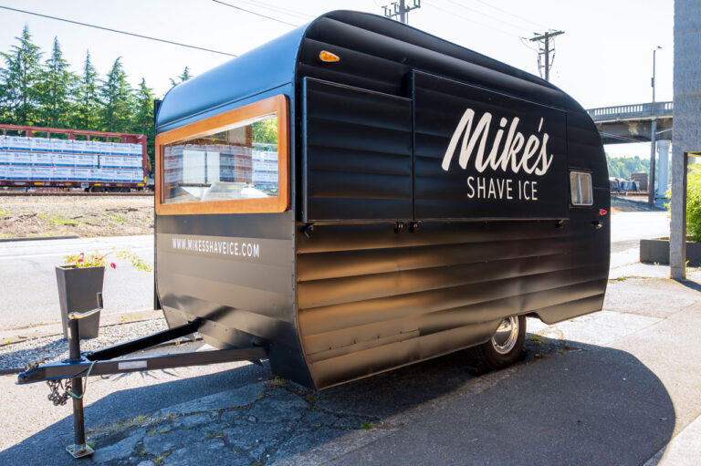 A black food trailer with "Mike's Shave Ice" written on the side is parked on a sunny street. The trailer has a serving window and is set on a concrete surface near greenery and a building structure.