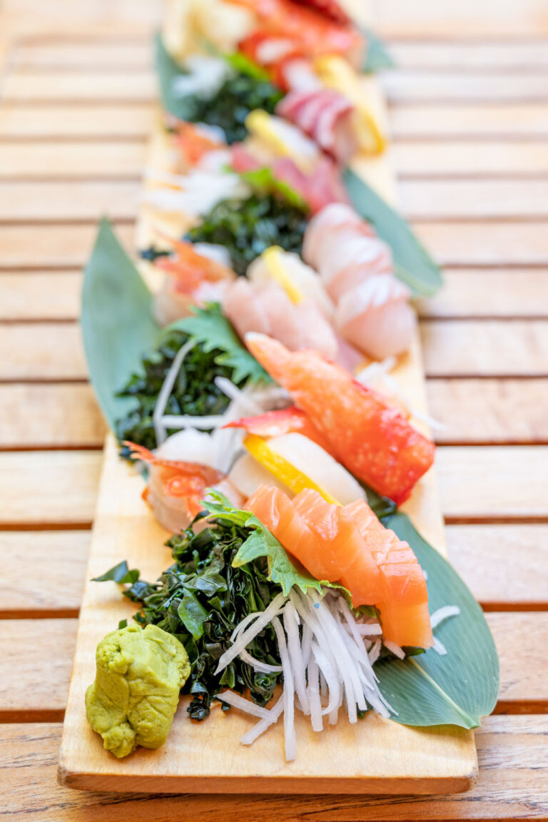 A wooden platter displaying an assortment of sushi pieces, including salmon, tuna, and shrimp, adorned with seaweed, daikon radish, and a dollop of wasabi. The presentation features green leaves as decoration.