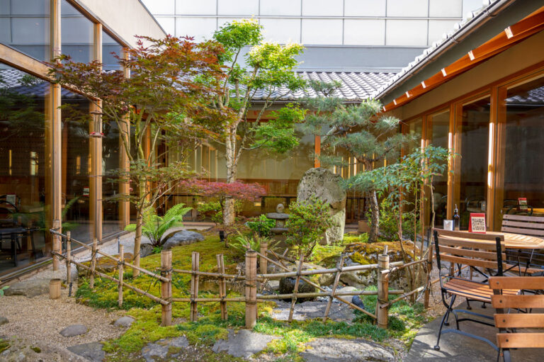 A tranquil Japanese garden enclosed by a wooden fence, featuring small trees, moss, rocks, and a stone lantern. Wooden tables and chairs sit nearby on a gravel path, surrounded by glass-walled buildings.