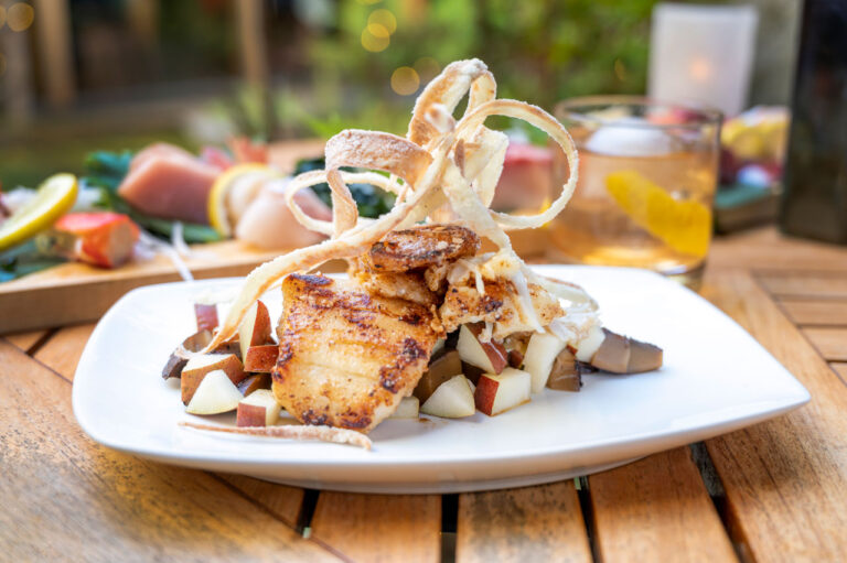 A gourmet dish featuring a seared scallop placed on a bed of diced pears and mushrooms, garnished with crispy onion strings, served on a white plate. A blurred wooden table setup with beverages and appetizers in the background.