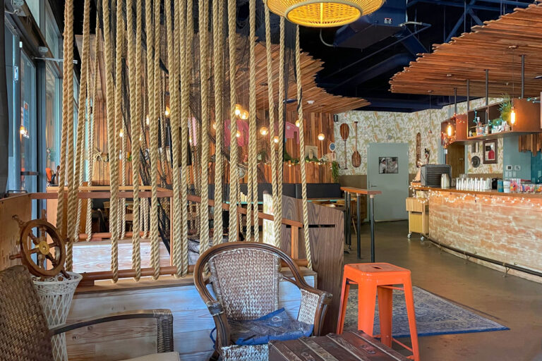 A cozy, rustic cafe interior featuring rope dividers, wooden tables, wicker chairs, and a bar area with string lights. The space has a warm and inviting atmosphere with wooden decor elements throughout.