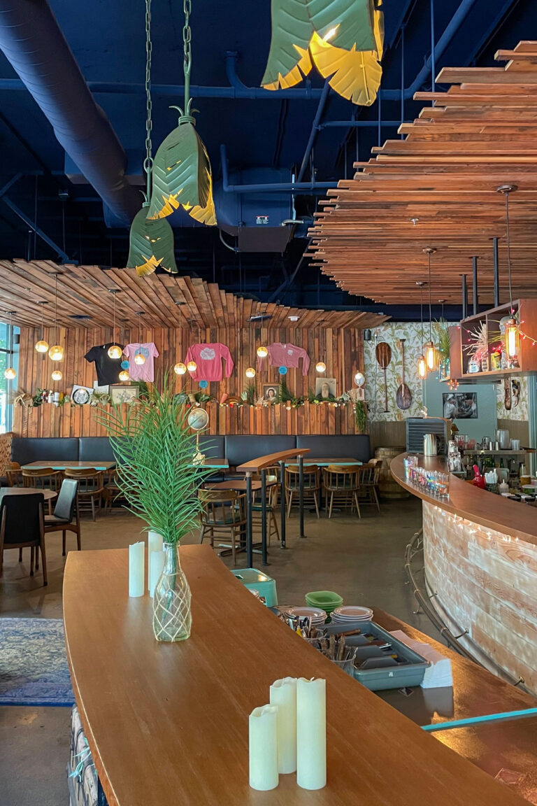 A stylish café interior with wooden accents and vibrant decor. Hanging plants and warm lighting create a cozy atmosphere. Tables and chairs are neatly arranged, with a vase and candles on the counter in the foreground.