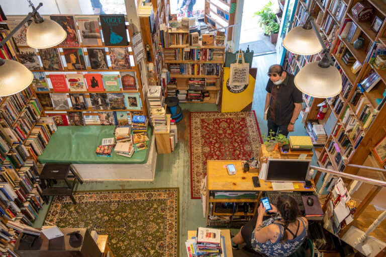Overhead view of a cozy bookstore with vibrant rugs, filled with books on shelves. A masked person stands browsing near the entrance, while another person sits behind the counter with a phone. Warm lighting creates a welcoming atmosphere.
