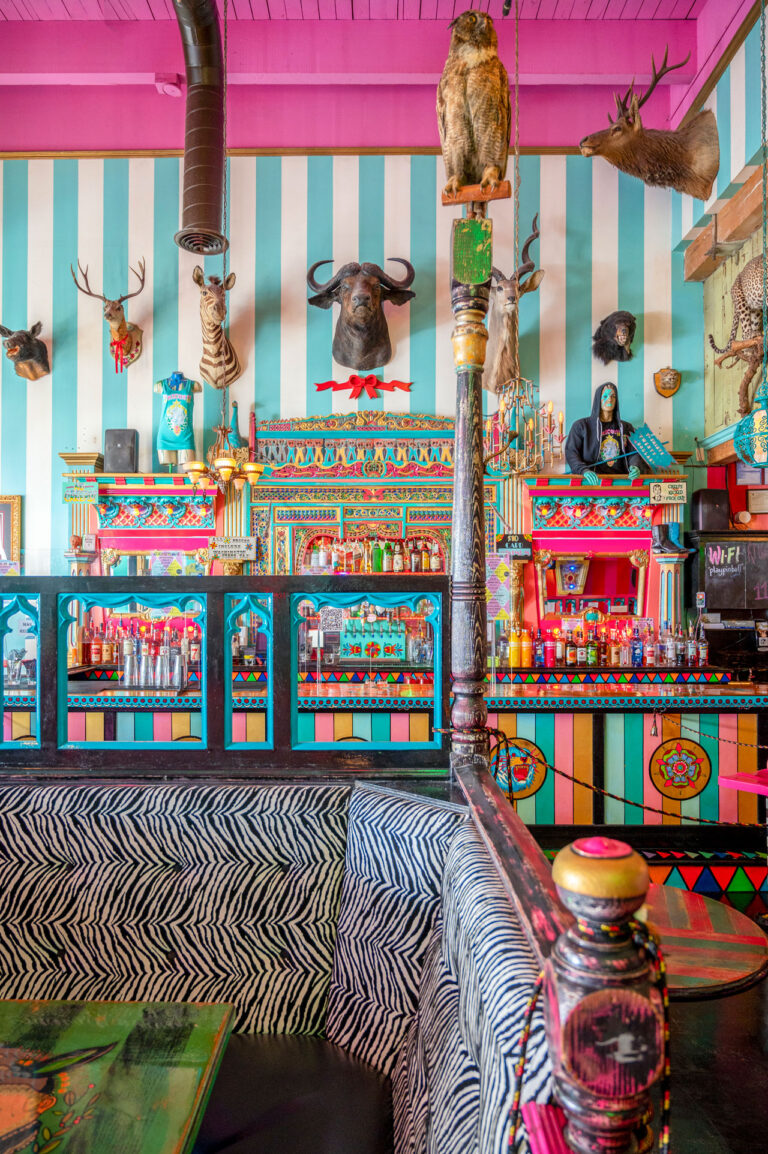 A vibrant, eclectic restaurant interior with colorful striped walls, taxidermy mounts, and decorative light fixtures. The bar is adorned with ornate patterns and surrounded by zebra-print seating.