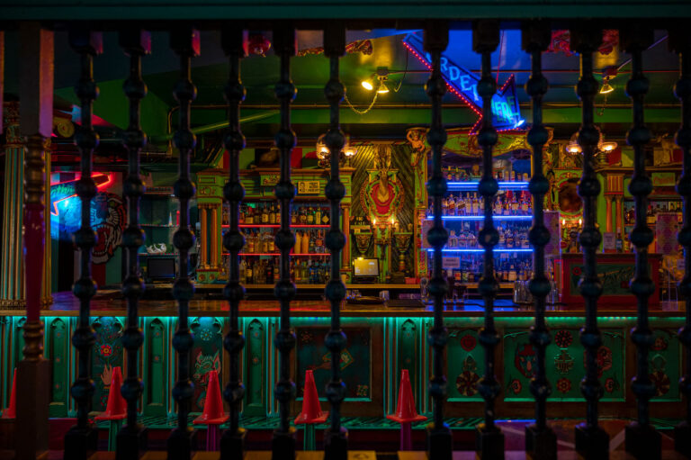 Dimly lit bar interior with colorful neon lights, decorated with ornate patterns and various bottles on shelves. A wooden railing with decorative spindles is in the foreground, partially obscuring the view of the bar.