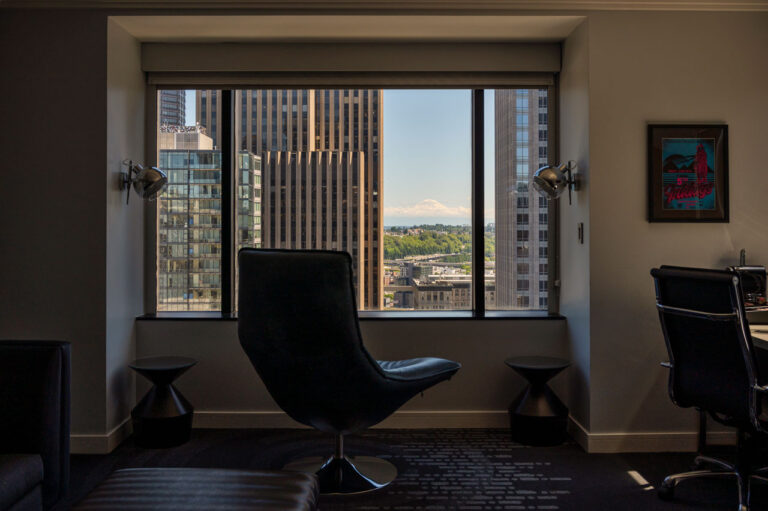 A modern office space with a swivel chair faces a large window, offering a view of tall city buildings and distant mountains under a clear blue sky. The room is decorated with contemporary lighting and a framed poster on the wall.