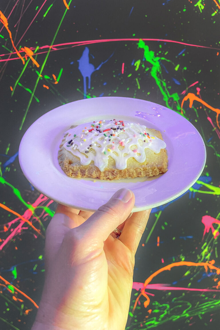 A hand is holding a white plate with a frosted pastry, topped with colorful sprinkles. The background features a vibrant, splattered paint design in neon colors.
