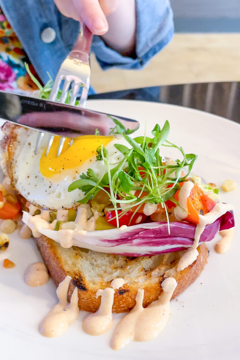 A person cuts into a slice of toast topped with a sunny-side-up egg, microgreens, radicchio, and vegetables. Drizzled sauce decorates the plate, and a colorful shirt partially visible beneath a denim jacket.