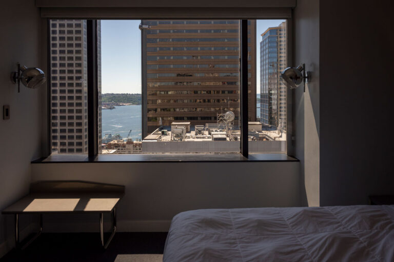 View from a hotel room window featuring city buildings and a distant water body under a clear blue sky. The room has a bed with white sheets and two wall-mounted lights. A small bench sits by the window.