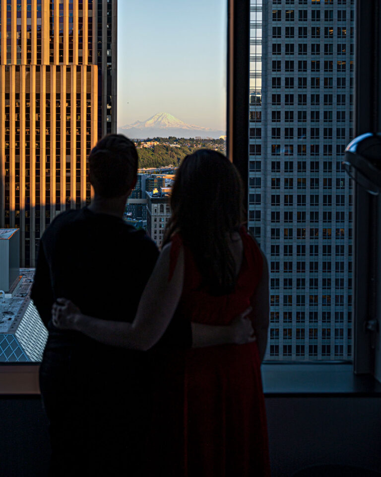 Two people stand by a window, embracing and looking out at a cityscape with tall buildings. A distant mountain is visible in the background, bathed in soft sunlight.