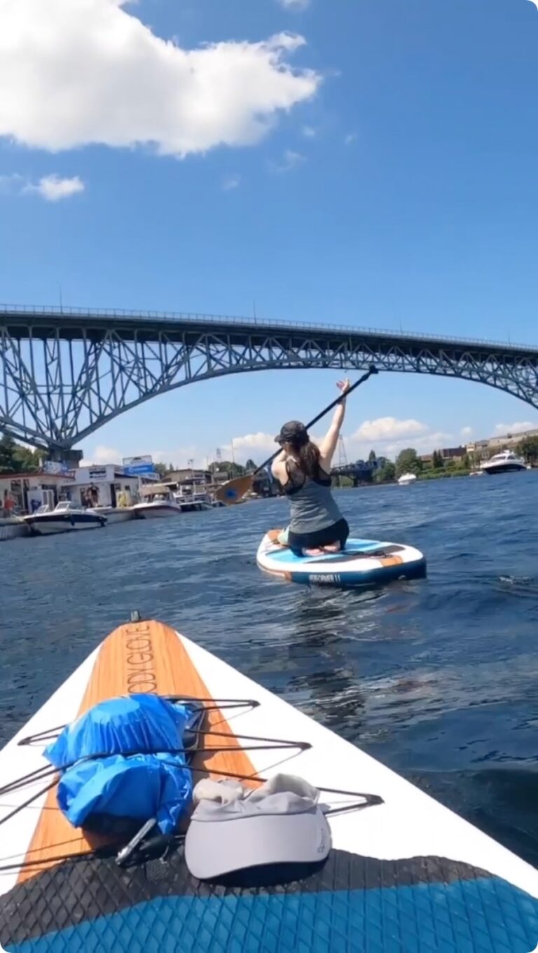 A person paddleboarding on a sunny day, raising their paddle triumphantly. They are on a river or lake with a bridge in the background. The foreground shows another paddleboard with a backpack and cap on it.